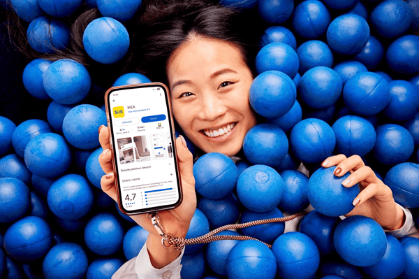 Uśmiechnięta kobieta zanurzona w basenie z niebieskimi piłkami trzyma w ręku smartfon z uruchomioną aplikacją IKEA widoczną na ekranie.
