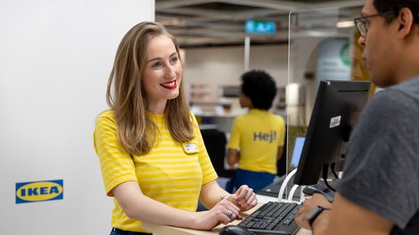 Uśmiechnięta pracowniczka IKEA rozmawia z klientem przy ladzie, na której znajduje się ekran komputera i klawiatura. 