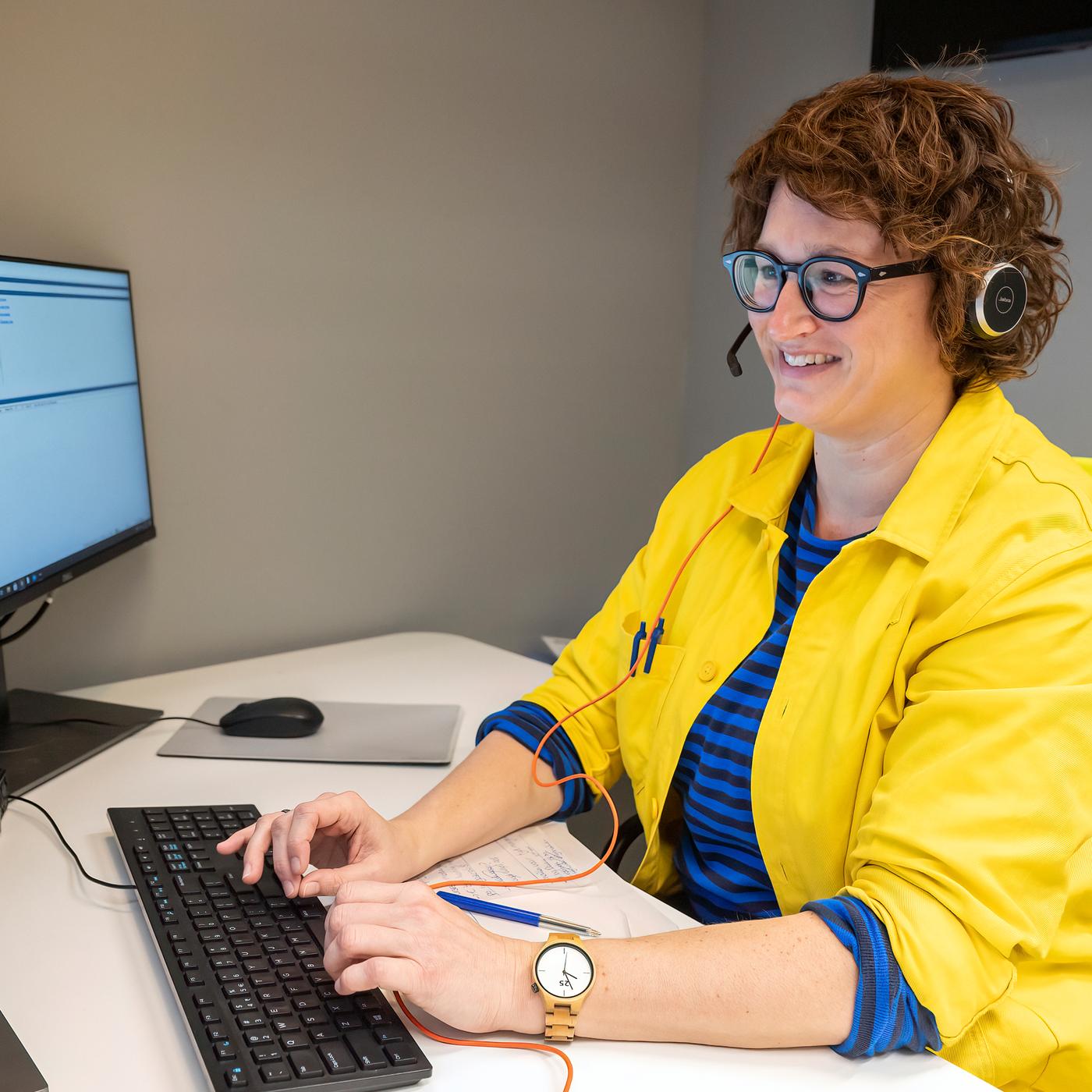 Usmievavá žena s okuliarmi a slúchadlami, oblečená v modro-žltej zamestnaneckej uniforme IKEA sedí pri stole a hľadí na monitor.