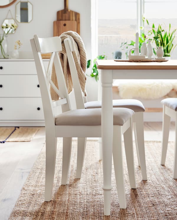 Ustawione na jutowym dywanie trzy białe krzesła i biały stół z dębowym blatem. W tle widoczny jest kredens z dwoma wazonami z kwiatami.