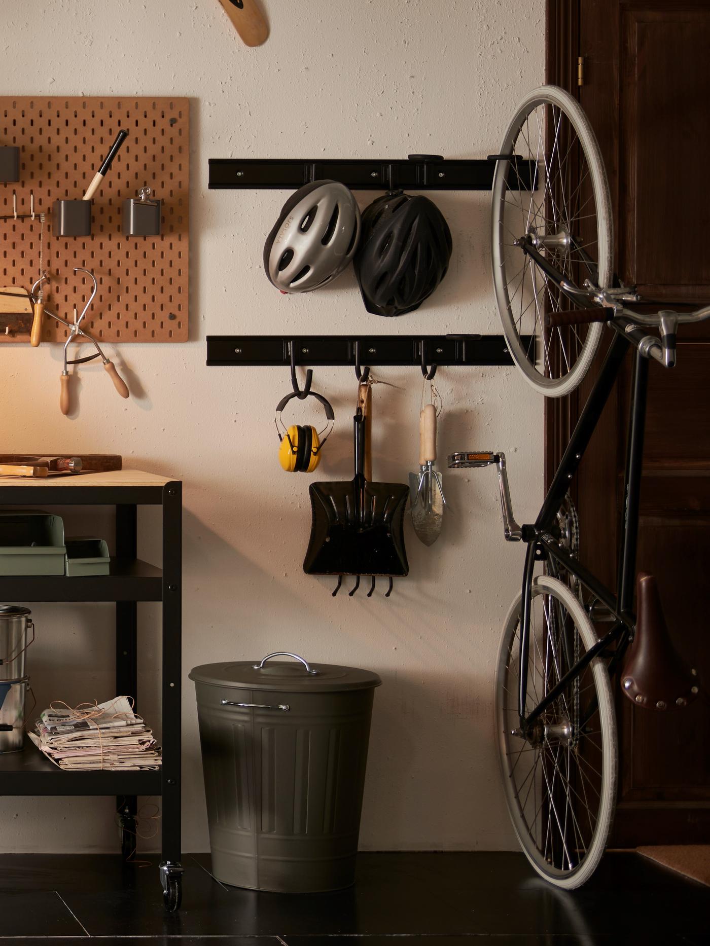 Ustawiony w garażu szary metalowy kosz z pokrywką i czarne metalowe wieszaki z powieszonymi na haczykach narzędziami ogrodniczymi.