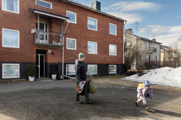 Utanför ett lägenhetshus går en man och mannens barn. I ena handen håller mannen två par skor och i andra handen en väska. Barnet har en liten ryggsäck på ryggen. 