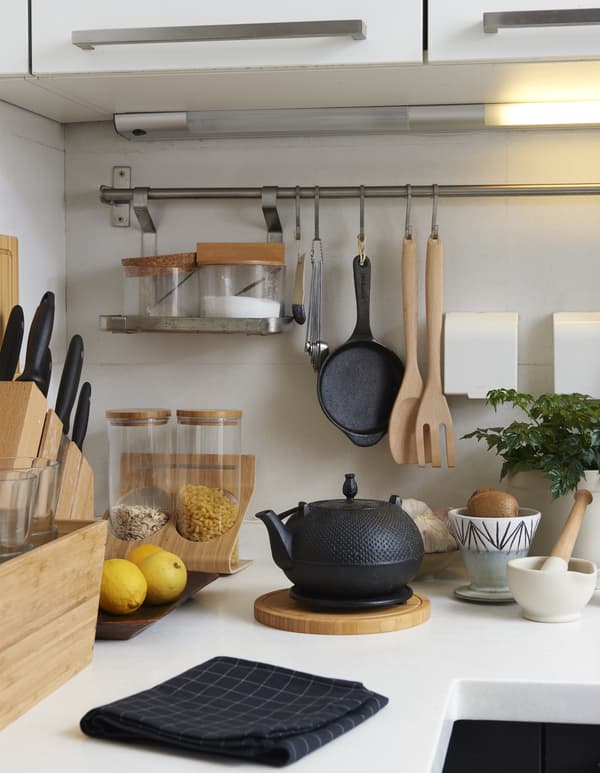 Utensili da cucina come coltelli, una teiera, uno strofinaccio nero e un tagliere con dei limoni su un piano di lavoro bianco.