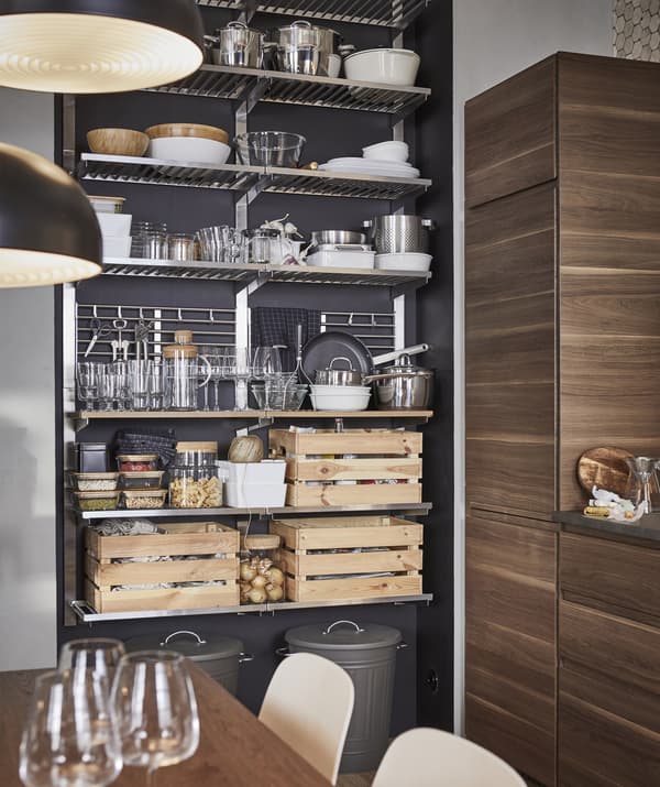 Utensilios de cocina, vajilla y cajas de madera almacenadas en un módulo de almacenaje abierto metalizado frente a una pared oscura.