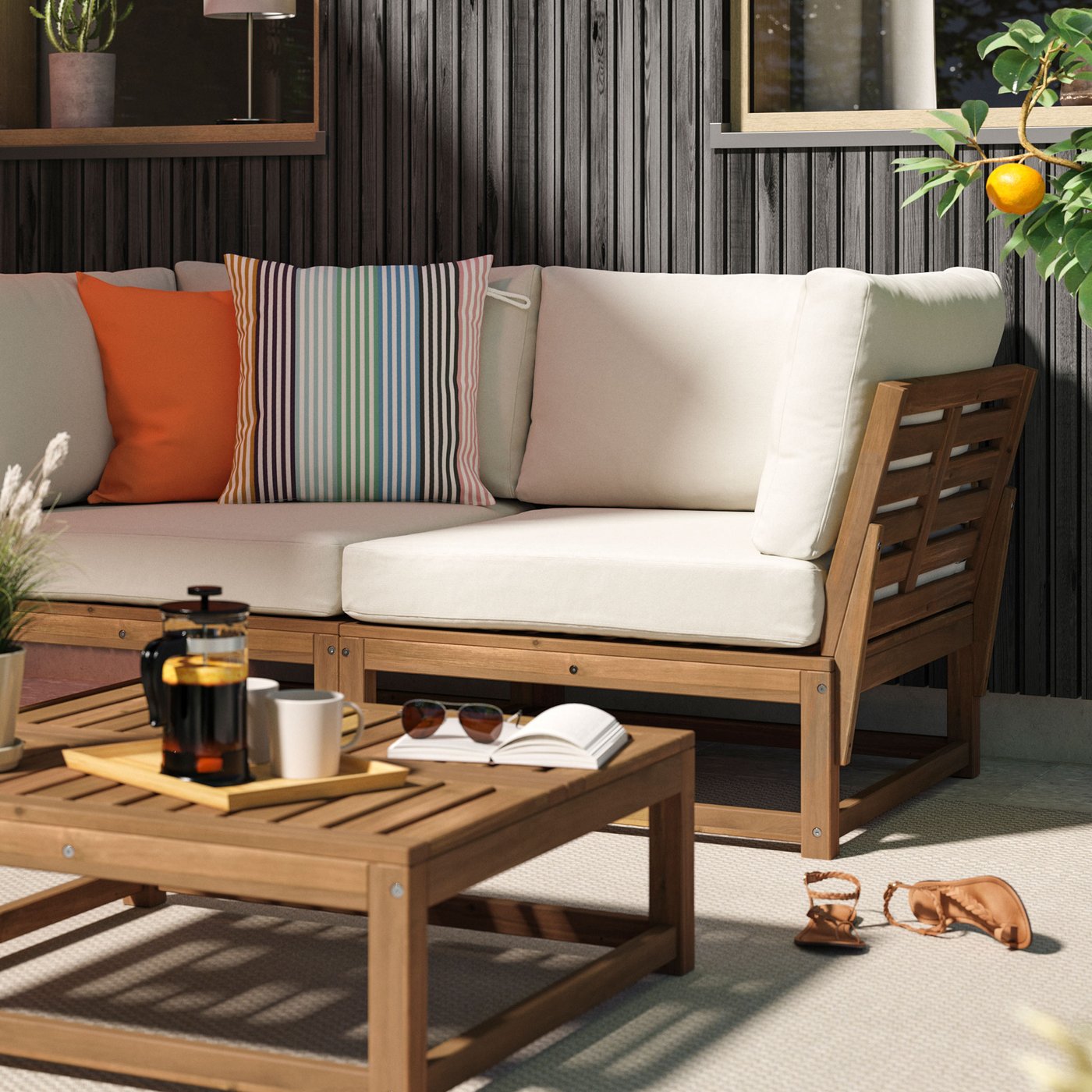 Uteplats i solen med en soffa i akacia med beige dynor och färgglada kuddar och matchande bord.