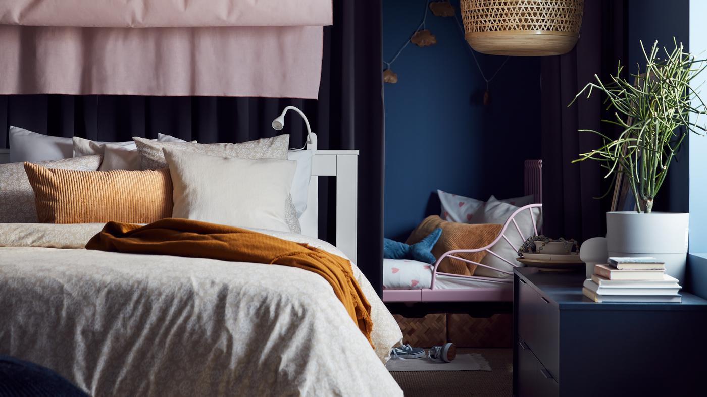 Uttrekkbar barneseng bak ei hvit dobbeltseng med beige og hvitt sengetøy og ei mørk beige pute, rosa gardiner og ei taklampe av bambus.