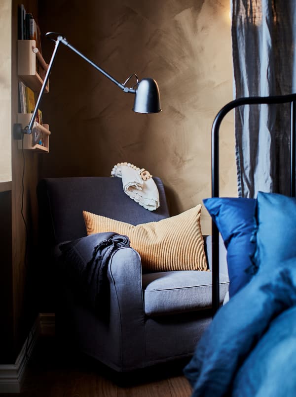Útulná spálňa s modrým kreslom GRÖNLID, svetlohnedým vankúšom, detskými knihami na stene a lampou na čítanie. 