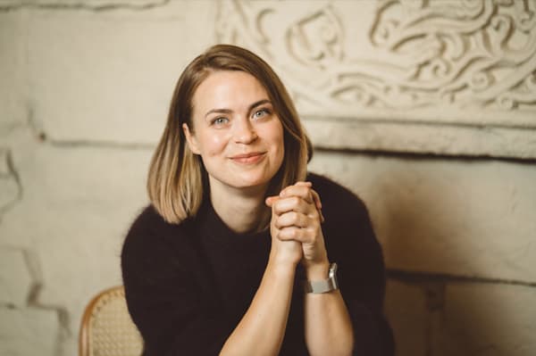 Uz gaišas akmens sienas fona redzama, melnā džemperī tērpusies, smaidīga sieviete vidēji gariem, brūniem matiem.