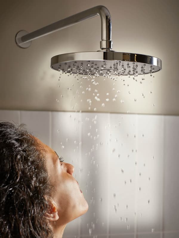 V bielo vykachličkovanej kúpeľni padajú kvapky zo sprchovej hlavice na tvár mladej ženy.