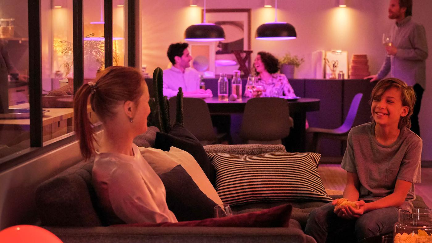V obývacej izbe sa žena na pohovke rozpráva s chlapcom, v pozadí sedí pri jedálenskom stole dvojica a nad ňou sú lustre NYMÅNE, ktoré svietia fialovým svetlom.