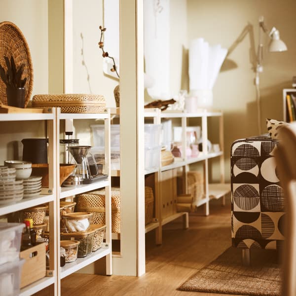 V obývacej izbe stoja pri stene otvorené úložné diely EKENABBEN a v nich misky GLADELIG, kanvica na kávu HÖGMODIG a škatule s vrchnákom.