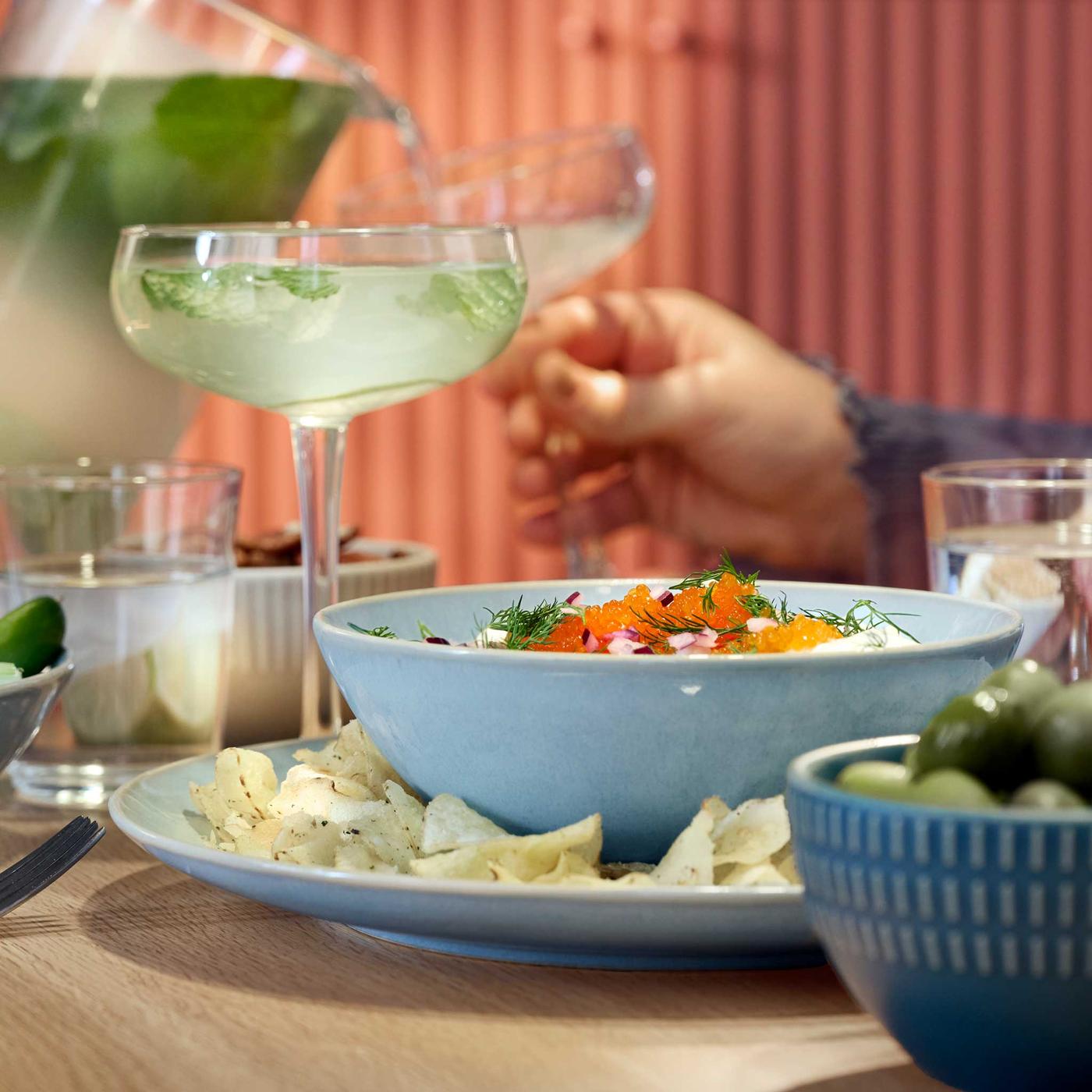 Vaaleansininen FÄRGKLAR-astiasto puisella pöydällä. Jalallisessa lasissa raikas juoma.