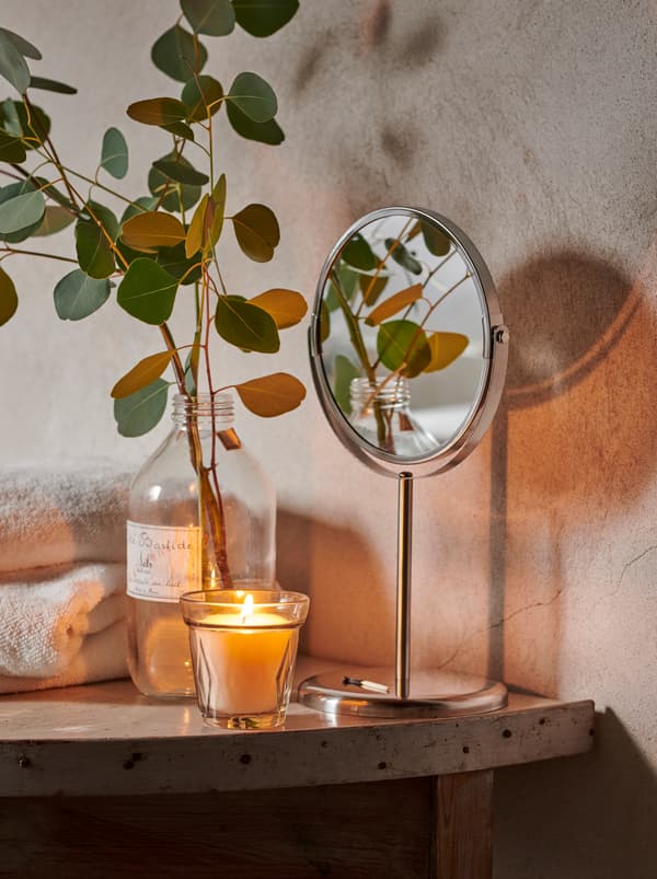 ผ้าเช็ดตัววางซ้อนกัน, เทียน VÄLDOFT/แวลดอฟต์ และขวดแก้วใส่่กิ่งและใบยูคาลิปตัส วางอยู่บนชั้นในห้องน้ำที่เรียบง่าย