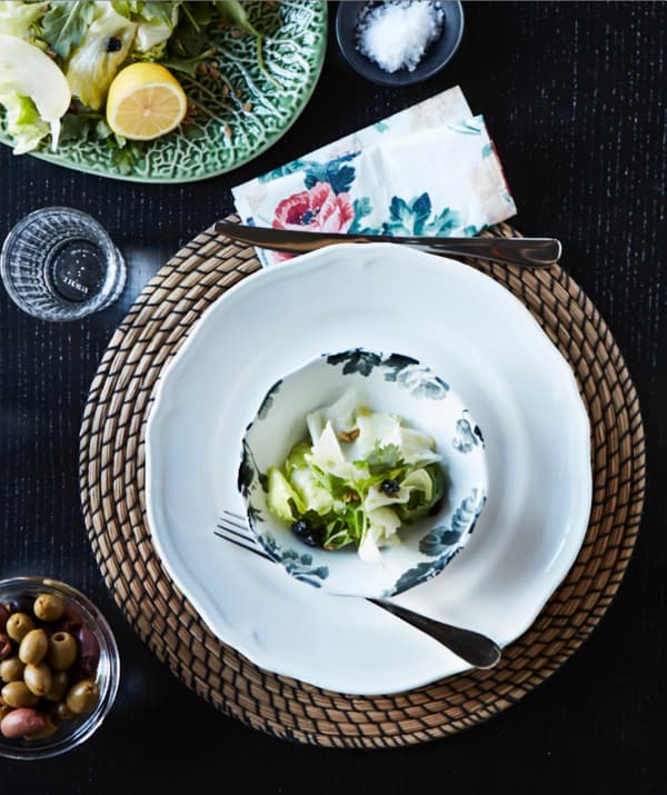 Vaisselle blanche et à motif floral déposée sur les napperons ronds en jonc de mer LÄTTAD sur une table à manger noire.