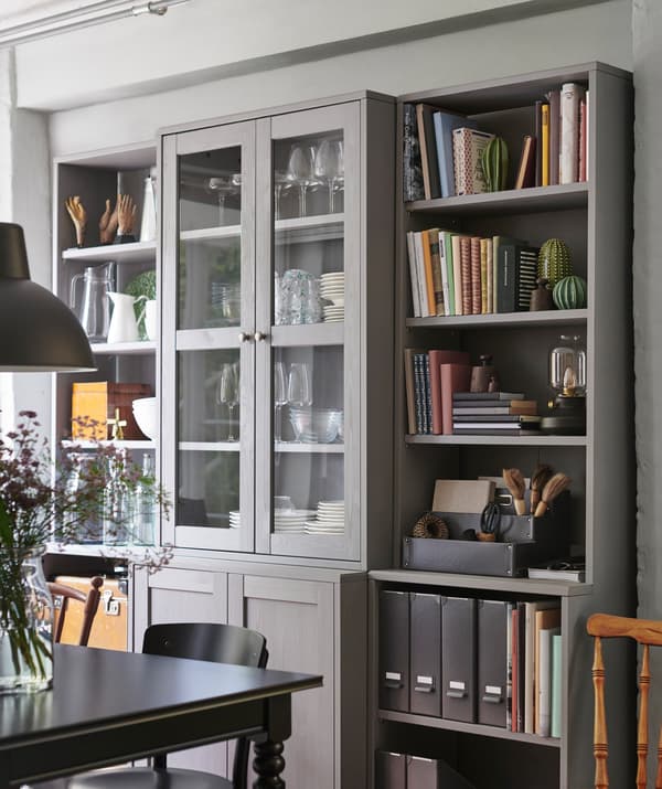 Vaisselle, livres et dossiers dans un meuble de rangement avec des étagères ouvertes et des armoires à portes vitrées à côté d’une table à manger.