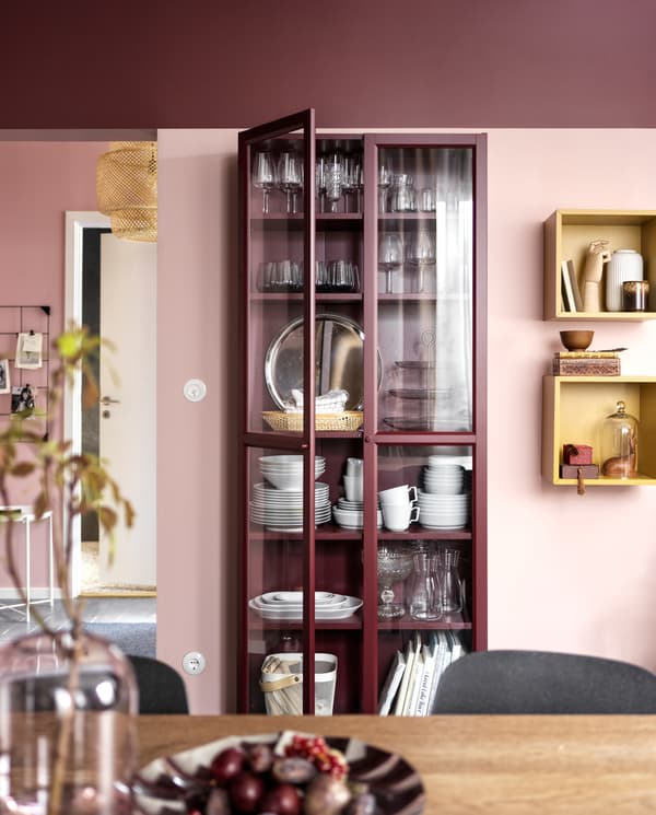 Vaisselle rangée dans une bibliothèque rouge foncé à portes vitrées. Deux cubes muraux ocre présentent des objets décoratifs.
