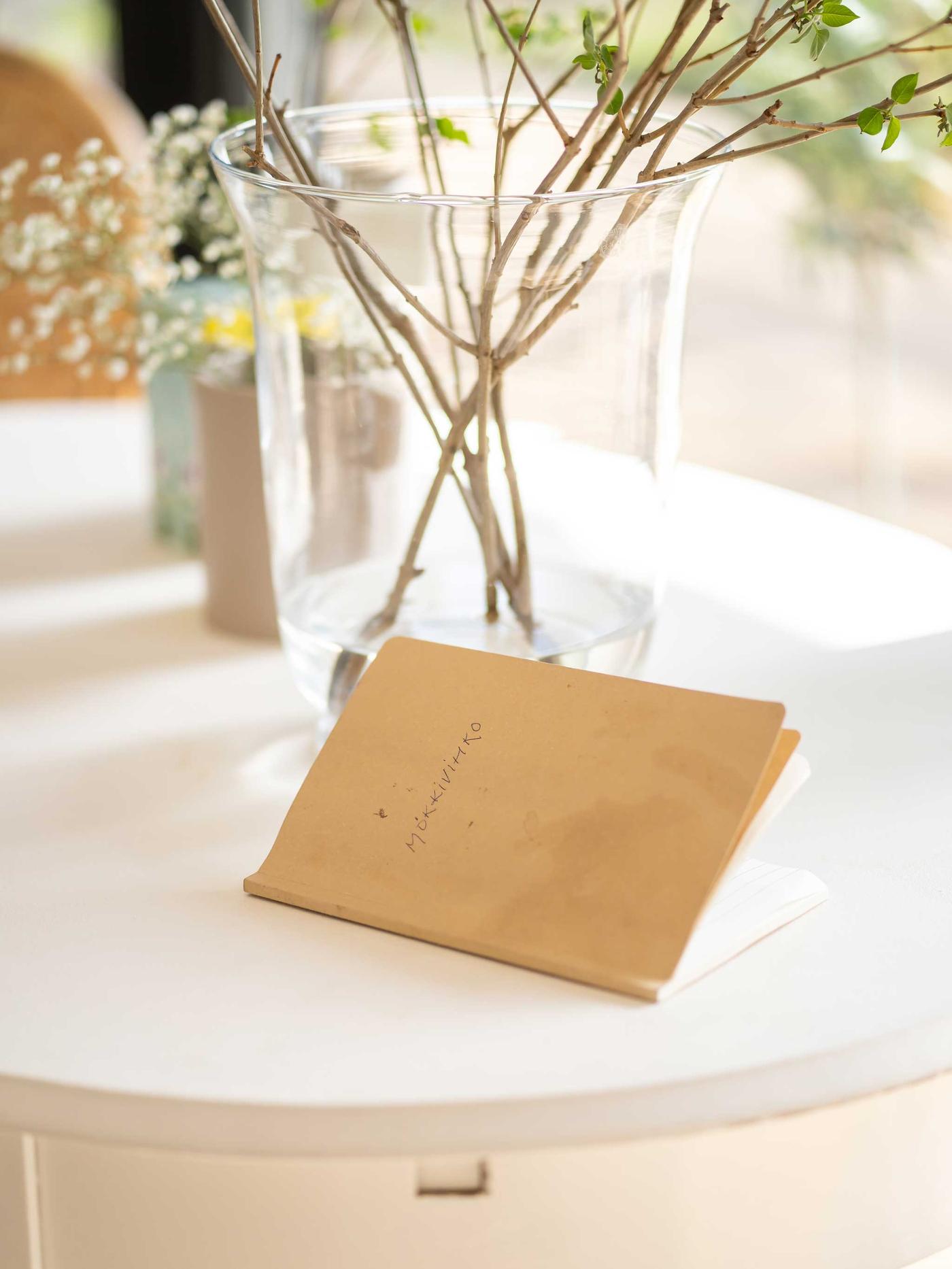 Valkoisella pöydällä on ruskea muistivihko ja luonnonkukilla täytetty maljakko.