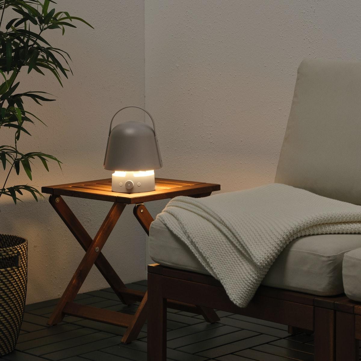 VAPPEBY lampe enceinte allumée et posée sur une petite table de jardin en bois à côté d'un fauteuil en bois avec un plaid blanc, le tout sur un petit balcon avec un mur blanc et un sol en caillebotis