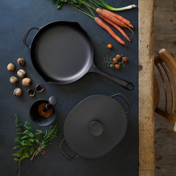 VARDAGEN casserole and frying pan are placed a black wooden table surrounded by cooking ingredients like carrots and nuts.