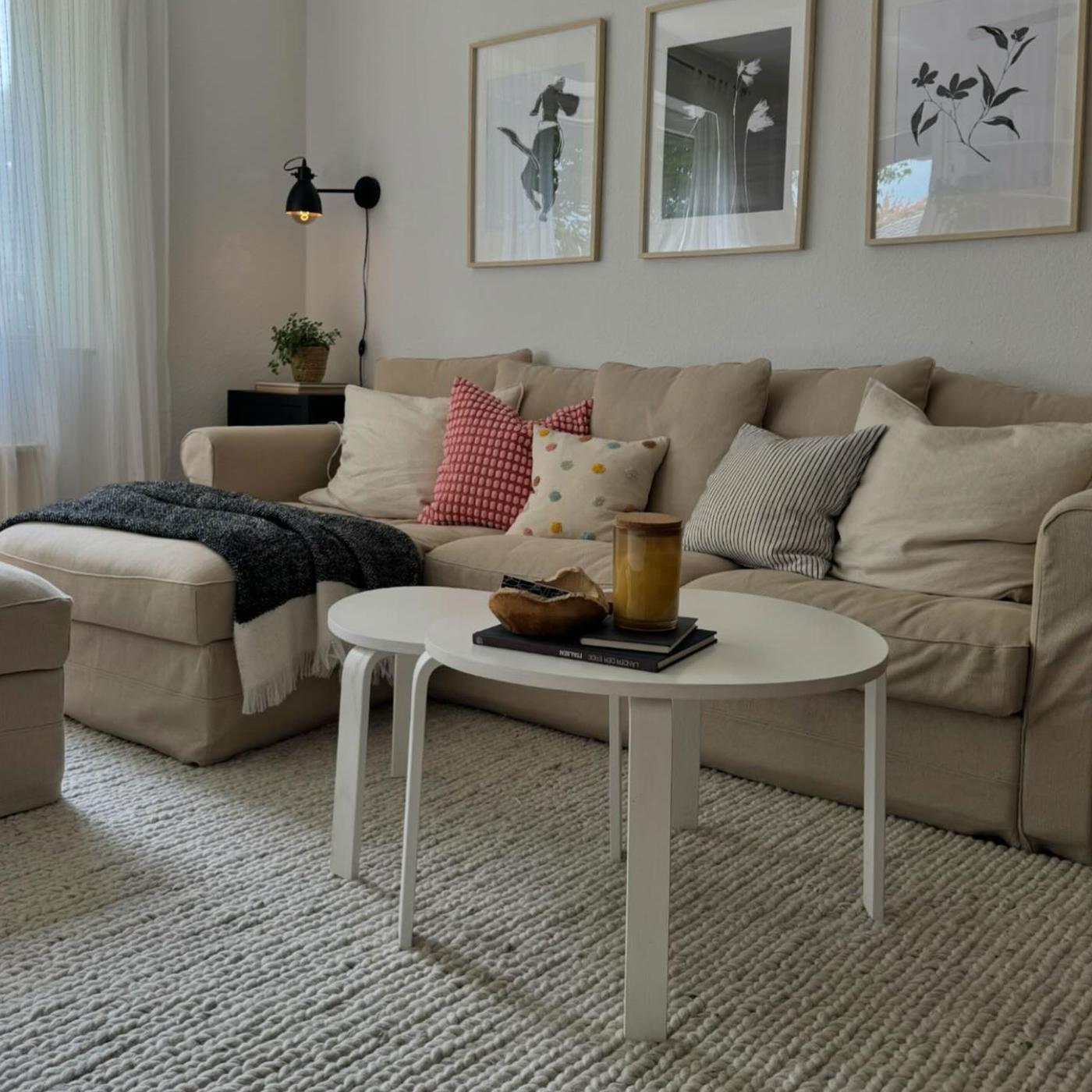 Vardargsrum med en beige 3-sitssoffa med schäslong och två vita soffbord i olika storlekar. Soffan är dekorerad med prydnadskuddar i färgerna rött, beige och vitt. Möblerna står på en texturerad matta i vitt. 