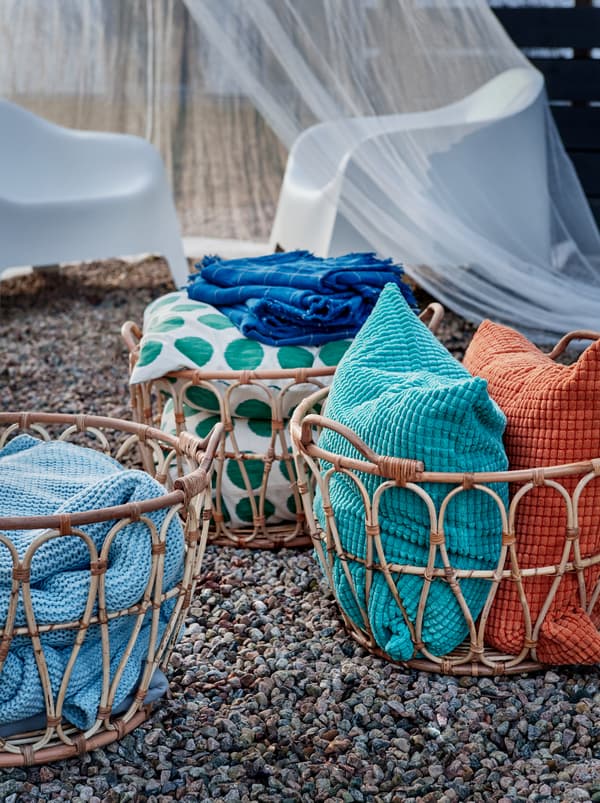 Vari cestini SNIDAD in rattan sulla ghiaia, contenenti plaid INGABRITTA, cuscini con fodera GULLKLOCKA e altri tessili.