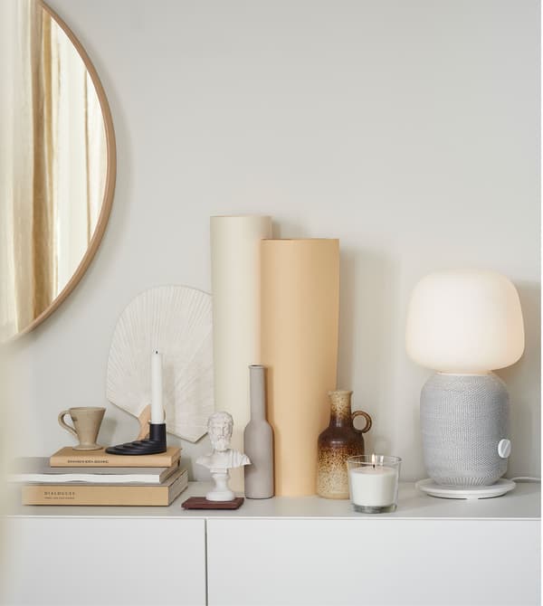 Varias decoracións de cores terrosas e unha lámpada de mesa gris e branca SYMFONISK sobre unha superficie branca.