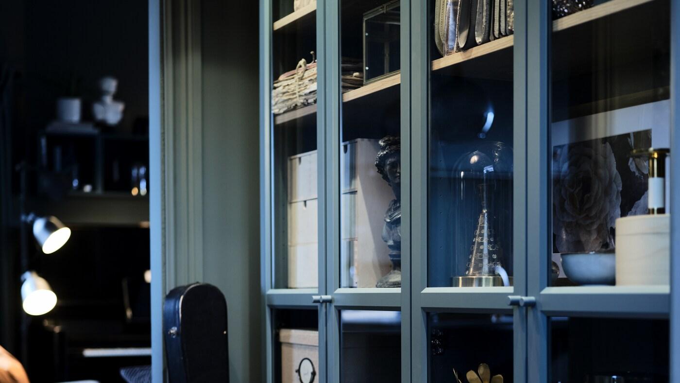 Varias librerías BILLY con puertas de vidrio dispuestas en paralelo con objetos en su interior. Dos lámparas iluminan la habitación en el fondo.