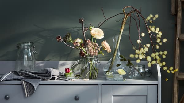 Varie composizioni di vasi e fiori su un buffet HAUGA grigio, una delle quali comprende una caraffa STORSINT - IKEA