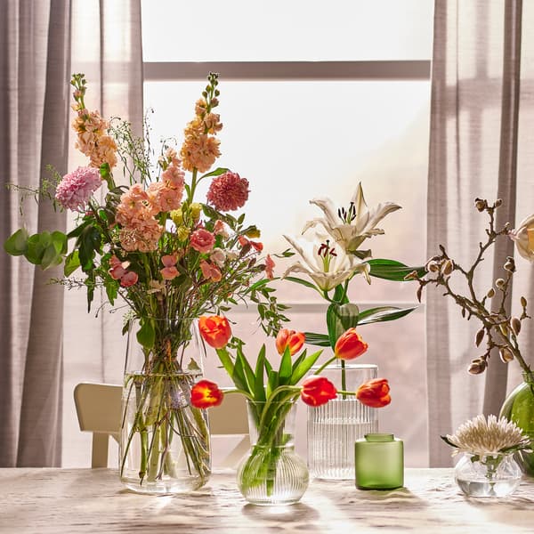 Varios arranxos de flores en vasos de vidro verde e vasos de vidro transparente sobre unha mesa á beira dunha fiestra con cortinas HILJA grises.