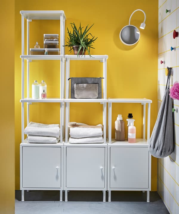 Varios estantes IKEA DYNAN brancos nun baño amarelo, usados para almacenar e colocar toallas e cosméticos.