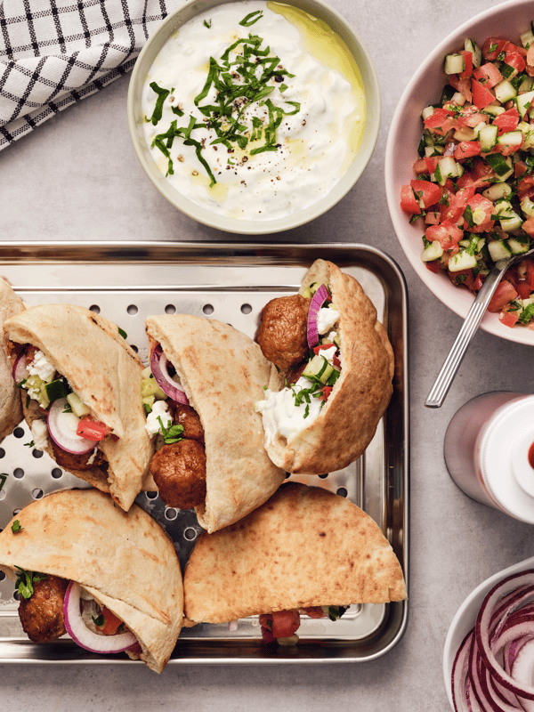 Varios panes de pita con albóndigas, verduras y salsa tzatziki en una bandeja de metal sobre una encimera de color gris claro.