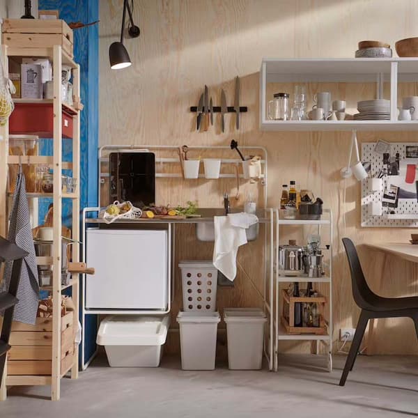 various shelves and storage units containing kitchen items.a towel hanging on its handle. Above there are hooks holding cups and other small items. To the right of the refrigerator, there are two white bins and a laundry basket with holes. 