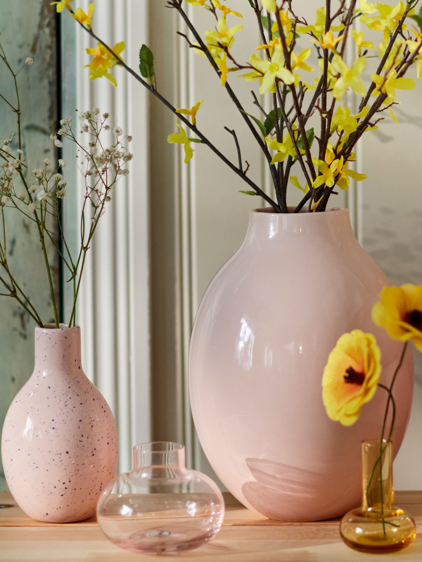 Vas i rosa keramik med gula blommor och dekorativa glasföremål på ett bord.