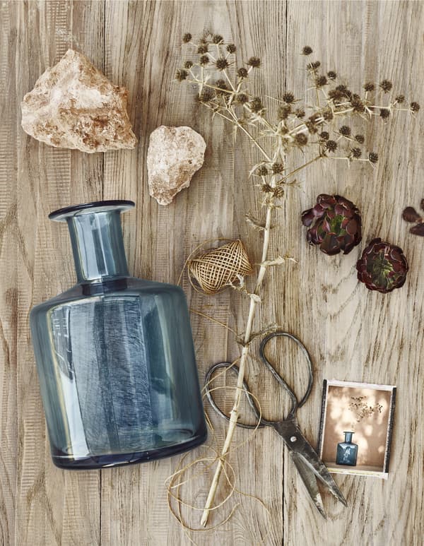 Vase en verre bleu, chardons séchés, ficelle, ciseaux et objets naturels disposés sur une surface en bois.