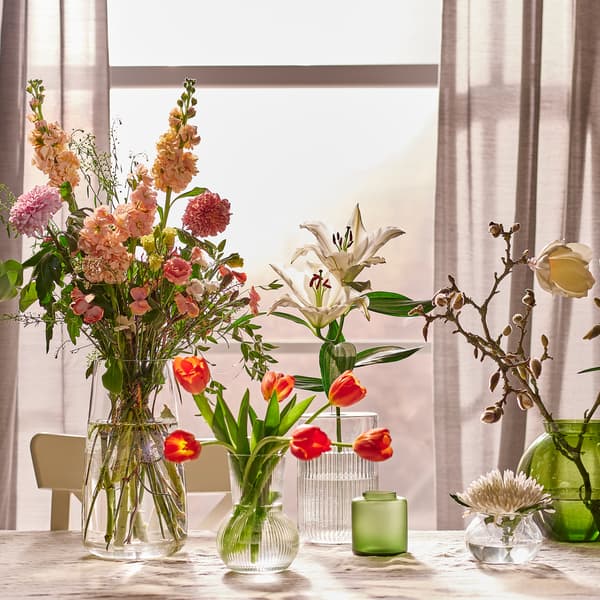 Vasi di diverse misure con fiori al loro interno, disposti su un tavolo con una tovaglia beige, di fronte a una finestra da cui entra la luce del sole.