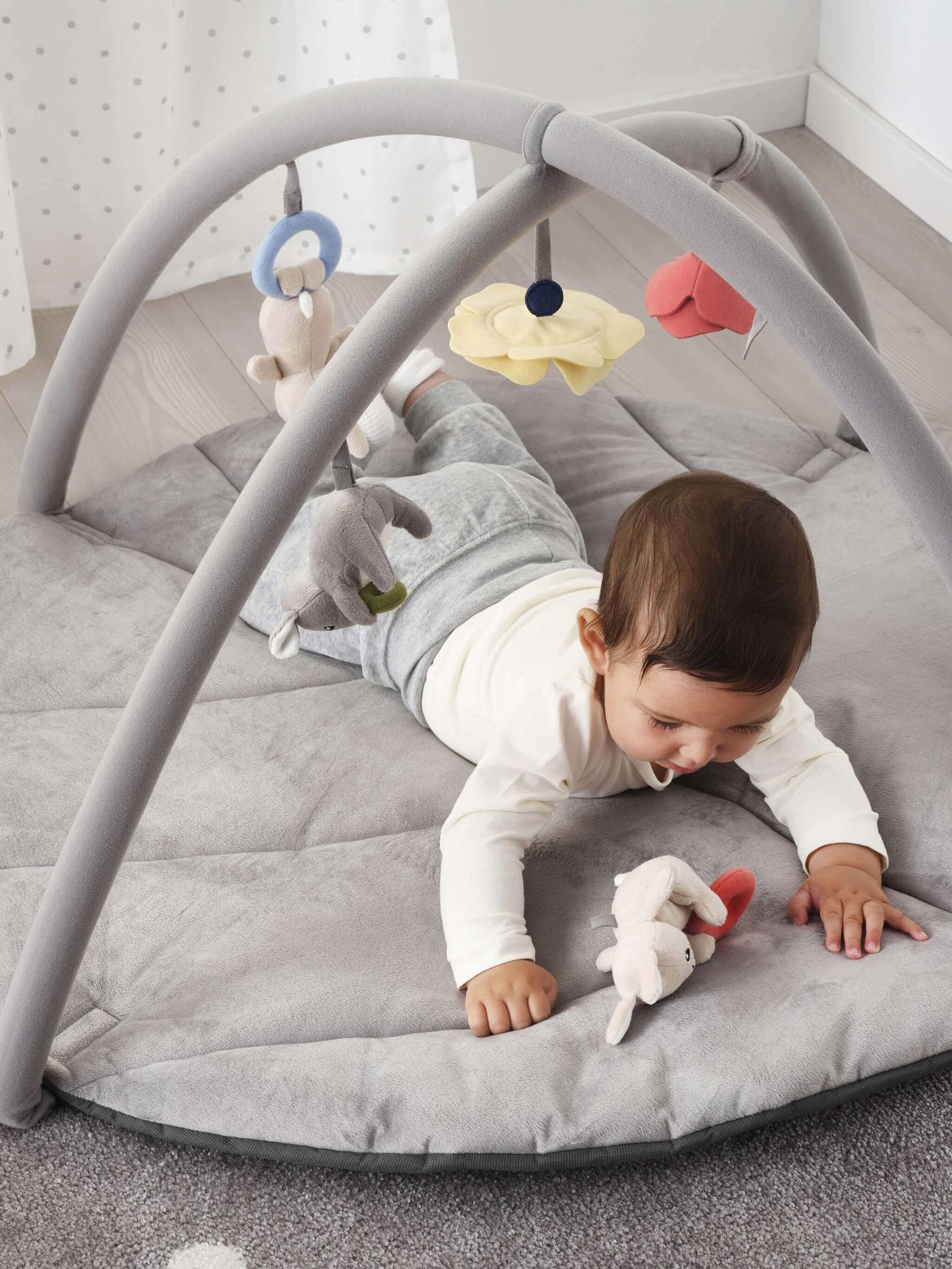 Vauva makaa harmaan GULLIGAST-jumppakaaren alla, josta roikkuu pehmoleluja.