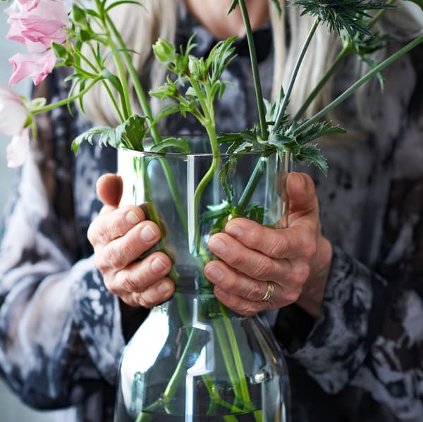 Váza OMTÄNKSAM s pekne naaranžovanými rastlinami, ktorú drží staršia žena. Vďaka svojmu tvaru sa váza dá jednoducho uchopiť a preniesť.