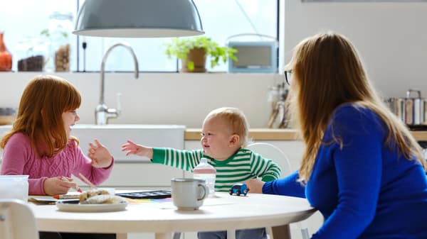 Ve světlé kuchyni sedí u stolu LISABO z jasanové dýhy dítě v židličce ANTILOP s dospělým a dalším dítětem.