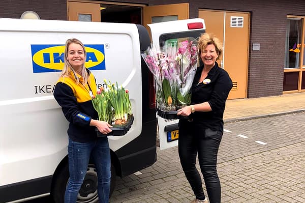 Vedle dodávky IKEA stojí dvě ženy, které drží v rukou rostliny orchidejí a tulipánů. Vypadají vesele.