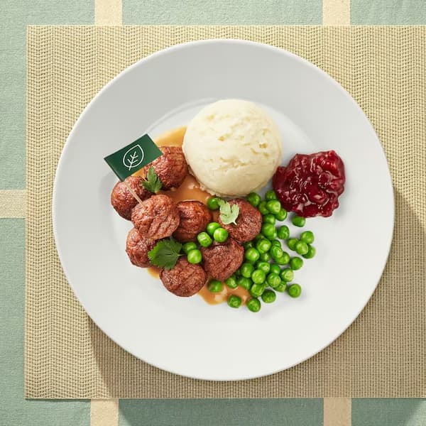 Veganské bezmasé kuličky s bramborovou kaší, omáčkou, hráškem a brusinkami na talíři. V jedné kuličce je zapíchnutá zelená vlajka na párátku s obrázkem listu.