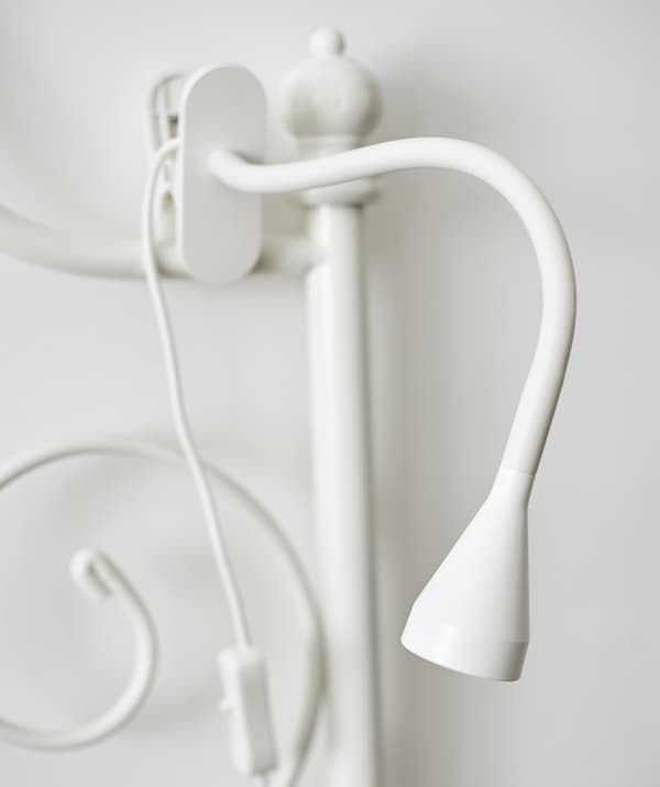 Veioză albă cu clamă cu cap lung îndoit, fixată de tăblia unui pat din fier forjat alb.