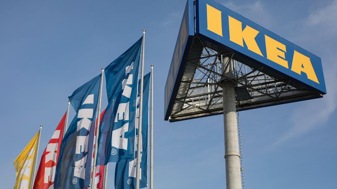 Veliki natpis IKEA na stupu, okružen plavim, crvenim i žutim zastavama s logotipom IKEA, na vedrom nebu.