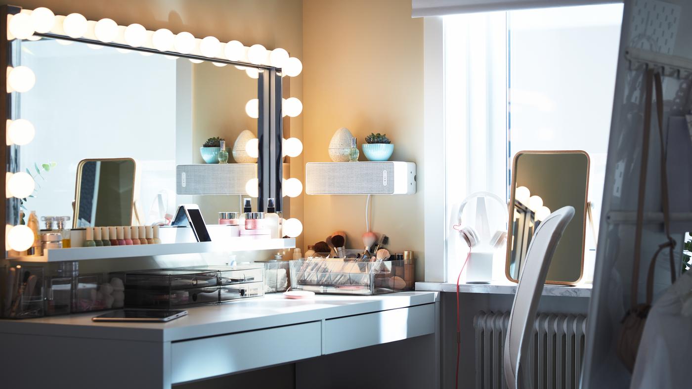 Veliko pravougaono ogledalo sa zidnim lampama iznad belog stola s mnogo prostora za odlaganje šminke, plus beli zvučnik na zidu.
