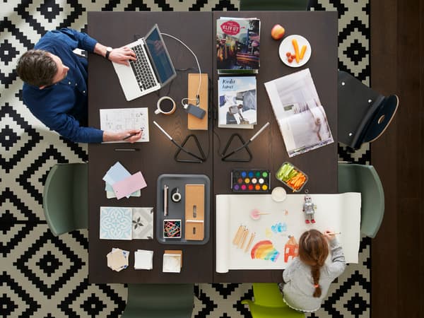 Velký stůl, z jedné strany sedí pracující muž s laptopem, z druhé strany je holčička, která si maluje.