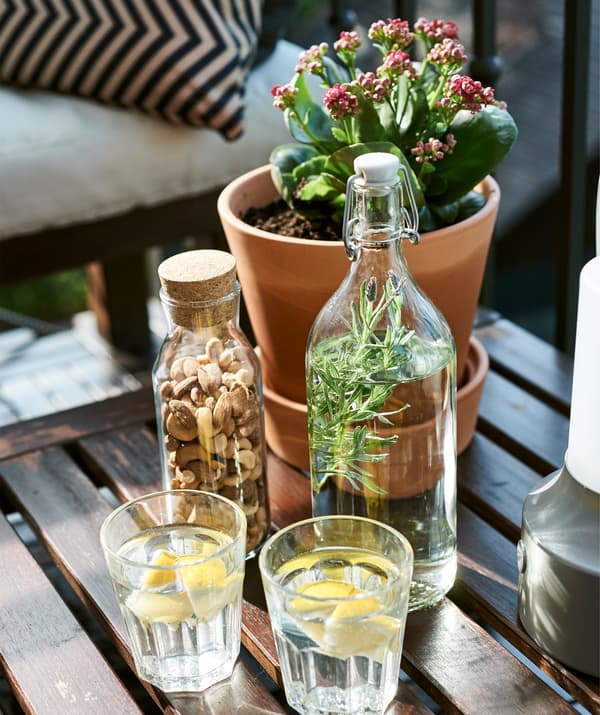Verres, carafe, bouteille et plante en pot sur une table basse d’extérieur en bois.