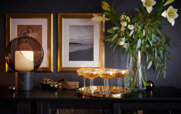 Verres de champagne sur un plateau doré, le tout posé sur une table noire. Une lampe EVEDAL et un vase de fleurs complètent la déco.