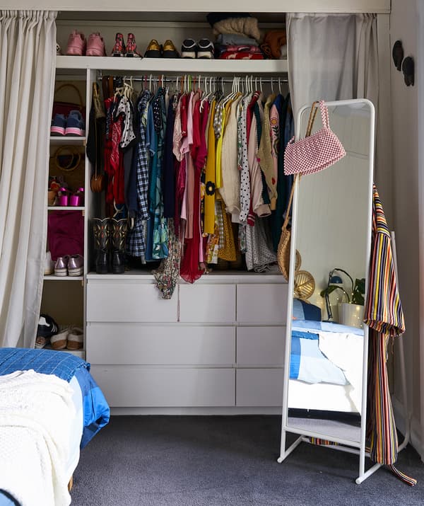 Vêtements sur cintres et chaussures sur étagères au-dessus de deux commodes, avec des rideaux de chaque côté et un miroir sur pied.