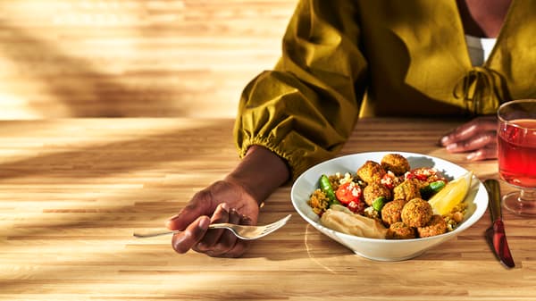 Vid ett träbord sitter en person med en gaffel i handen. Framför personen står en tallrik med couscous och falafel. Ett dricksglas står bredvid.