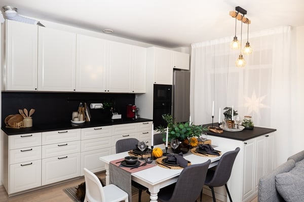 Вид на рабочую зону кухни с плитой, духовкой и бытовой техникой; рядом — обеденный стол и окно со шторами.