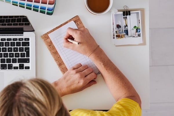 Вид сверху: дизайнер интерьеров IKEA в желтой форме сидит за столом с компьютером, кружкой чая, палитрой и картинками для вдохновения и делает заметки на бумаге.